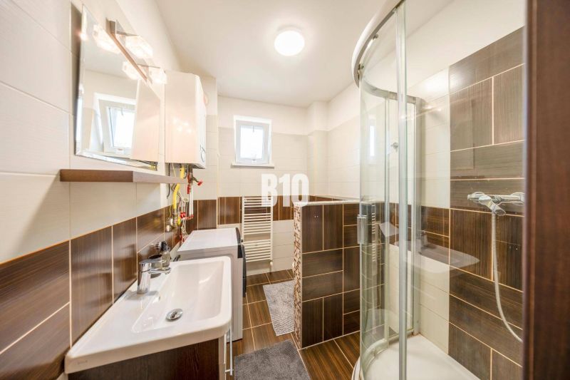 ŠTARTOVACÍ 2-IZBOVÝ BYT S KLIMATIZÁCIOU,  BALKÓNOM, PARKOVACÍM STÁTÍM  - Kúpeľňa v 2-izbovom byte s tmavými obkladmi a sprchovacím kútom.
