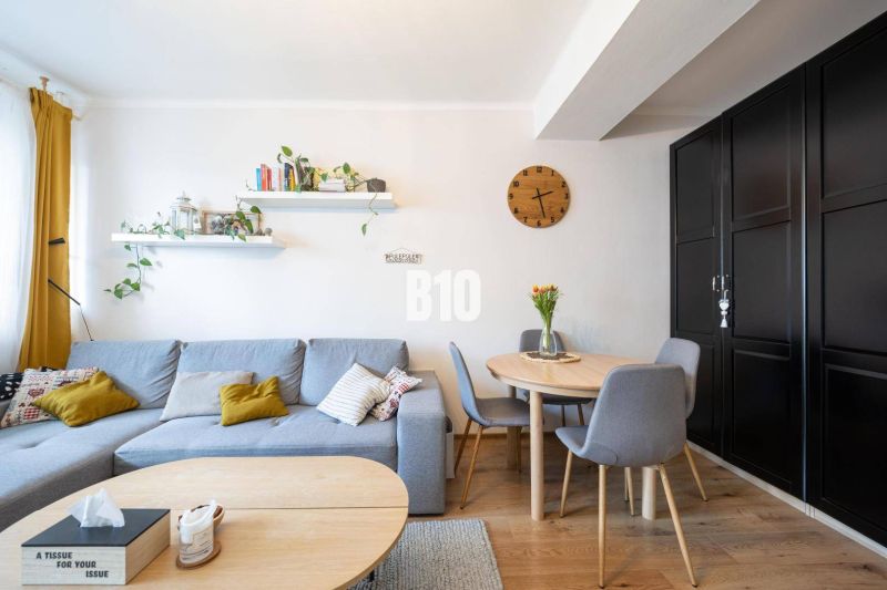 2-izbový byt po rekonštrukcii | Vietnamská ulica, Ružinov | Loggia | B - Obývačka so sivou sedačkou, okrúhlym stolom a podlahou s dreveným dekorom v 2-izbovom byte.