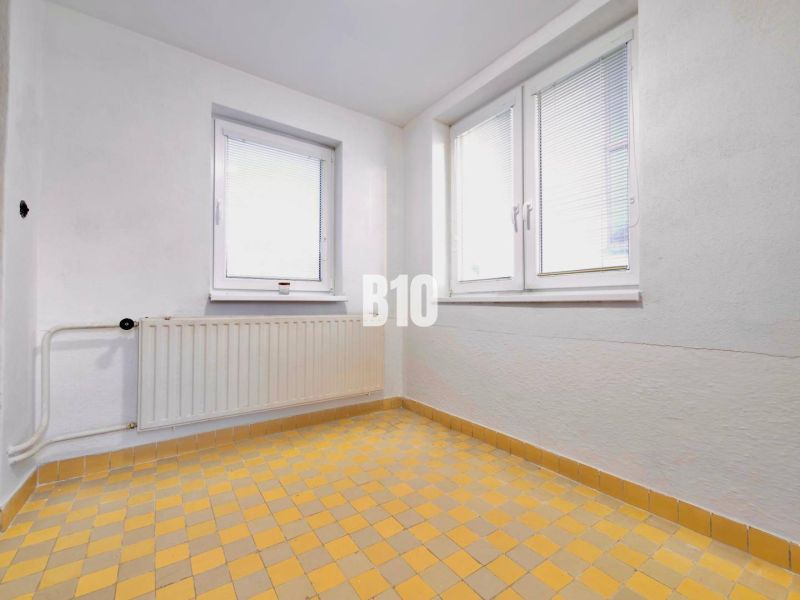 Rodinný dom s rovinatým 10a pozemkom - Biela stena, okná, radiator a žltá kachľová podlaha v rodinnom dome.