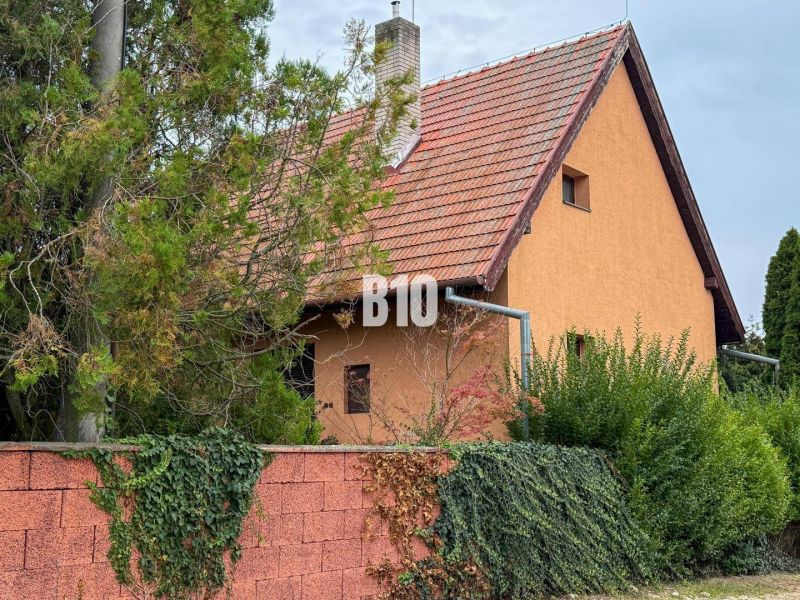 Priestranný rodinný dom s garážou na veľkom pozemku v obci Vlkas - Rodinný dom vo Vlkase s červenou strechou obklopený zeleňou a tehlovým plotom.