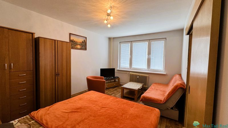 Štartovací byt s balkónom a výhľadom, ktorý vás bude baviť! - Izba s nábytkom v 1-izbovom byte, podlaha s dreveným dekorom, oranžové poťahy na sedačke.