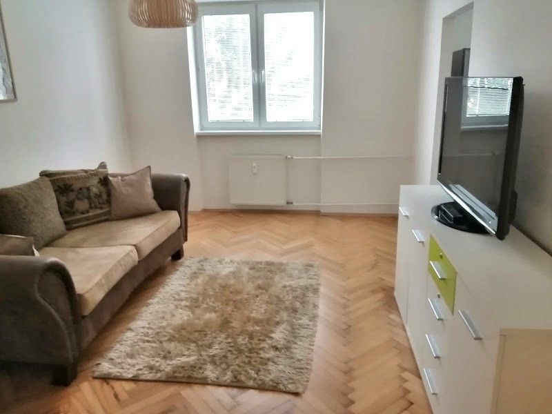 PRENÁJOM: ÚTULNÝ 2 IZBOVÝ BYT S BALKÓNOM, UHLISKO - Obývacia izba v 2-izbovom byte s pohodlnou sedačkou, televízorom a podlahou s dreveným dekorom.