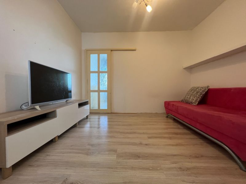Na predaj 1-izbový byt s loggiou - Žiar nad Hronom, M. Chrasteka 1B718 - Obývacia izba s TV, červenou sedačkou a podlahou s dreveným dekorom v 1-izbovom byte.