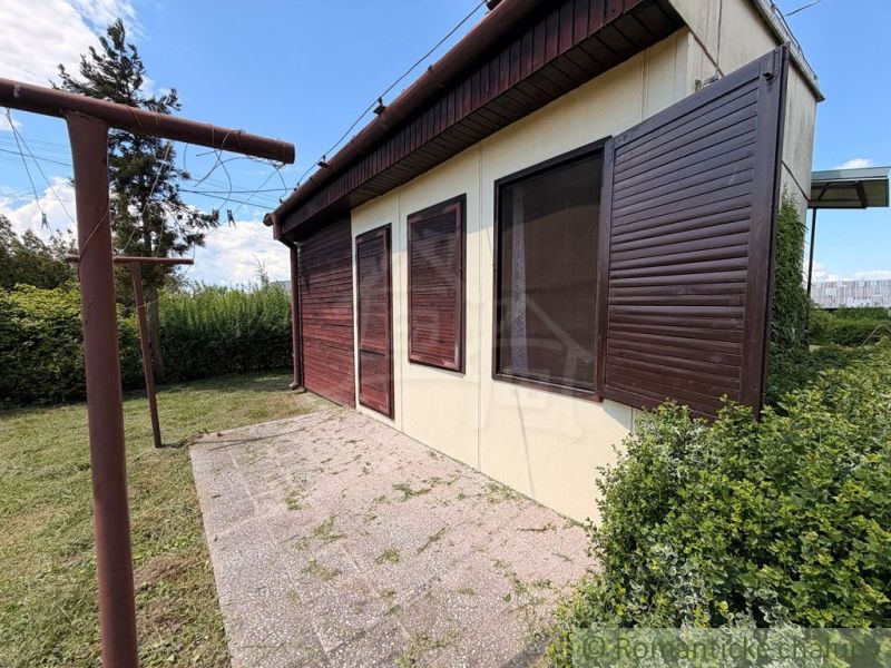AREÁL 3 REKREAČNÝCH CHÁT PRI PODHÁJSKEJ – IDEÁLNE PRE DETSKÉ TÁBORY, R - Chata v Nových Zámkoch so zatvorenými drevenými okenicami a okolitou zeleňou na záhrade.