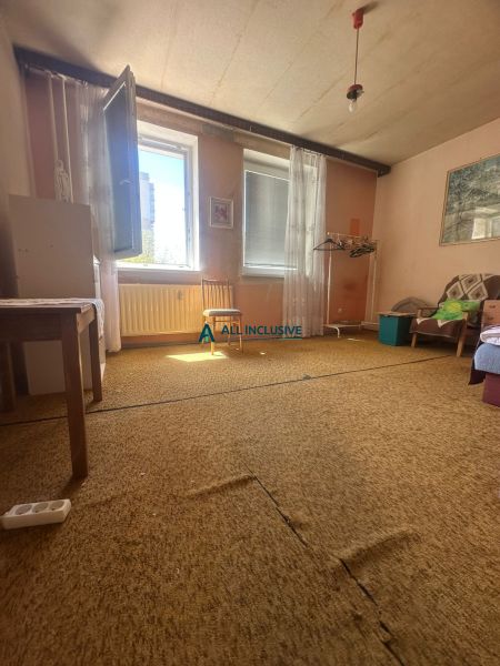 Rezervovaný - Na predaj priestranný 2-izbový byt v Topoľčanoch - Izba v 2-izbovom byte s kobercom, otvoreným oknom a jednoduchým nábytkom.