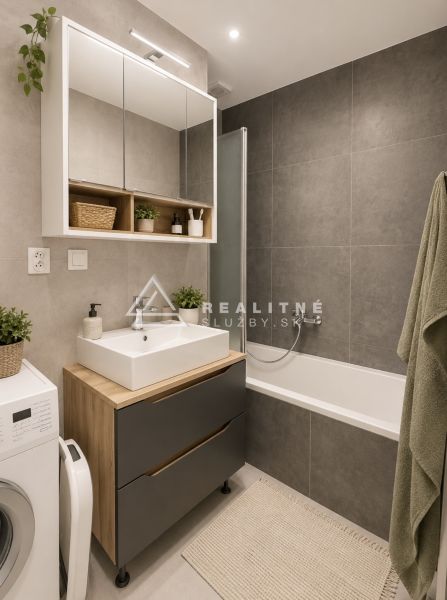 NOVOSTAVBA , BAŠTOVÁ 48 M2 2-IZBOVÝ BYT S LOGGIOU  - Kúpeľňa v 2-izbovom byte s vaňou, skrinkami a práčkou, šedé dlaždice.