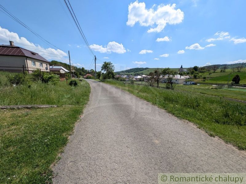 Slnečný stavebný pozemok 1 186 m² - Karná | možnosť dokúpiť 1 ha - Cesta obklopená trávou vedie cez vidiecku oblasť v Karnej s výhľadom na domy.