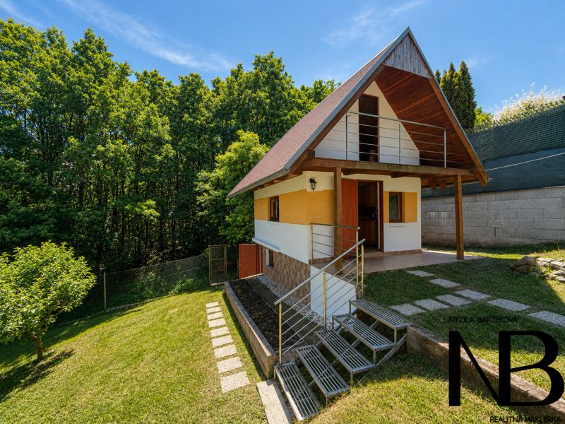 Záhrada s murovanou chatkou a výhľadom, Považská Bystrica -Klučina - Chata s dreveným dekorom v Považskej Bystrici na Klučine, obklopená zeleňou a stromami.