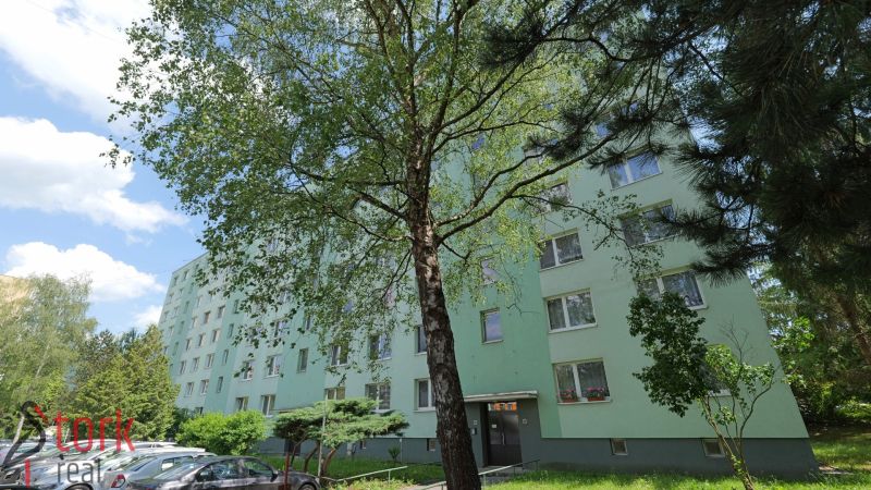 Na predaj - 3i BYT s veľkorysou výmerou - centrum PB - Považská Bystrica, Štúrova: Panelový dom obklopený stromami a zaparkovanými autami na ulici.