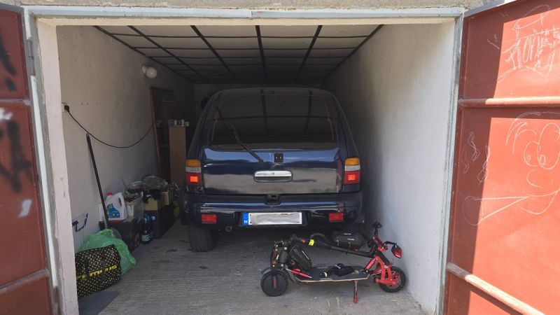 NA PREDAJ MUROVANÁ GARÁŽ 18 m² VO ZVOLENE – ELEKTRINA, VLASTNÝ POZEMOK - Garáž s autom a elektrickou kolobežkou, uloženými predmetmi a kresbou na dverách.