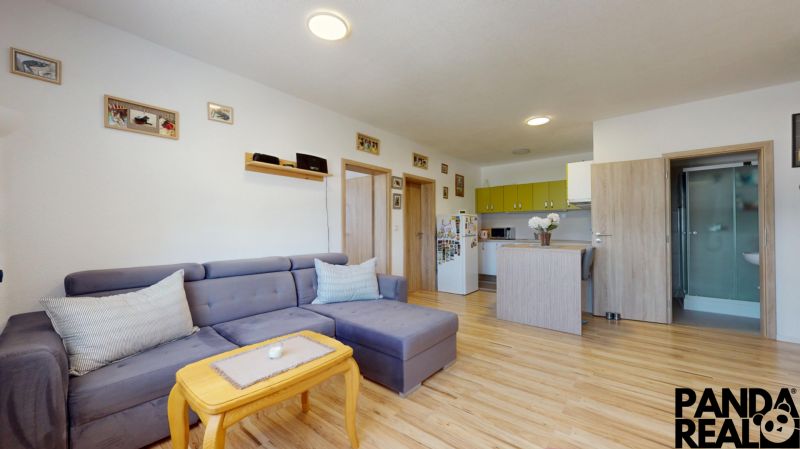 Moderný 3-izbový byt s balkónom a 2 parkovacími miestami - Ludanice - Obývacia izba s rohovou sedačkou a kuchyňou, podlaha s dreveným dekorom.