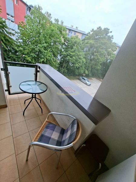 Priestranná garzónka s balkónom v novostavbe. - Balkón garsónky s výhľadom na zeleň a parkovisko, miestnosť so stolom a stoličkou.
