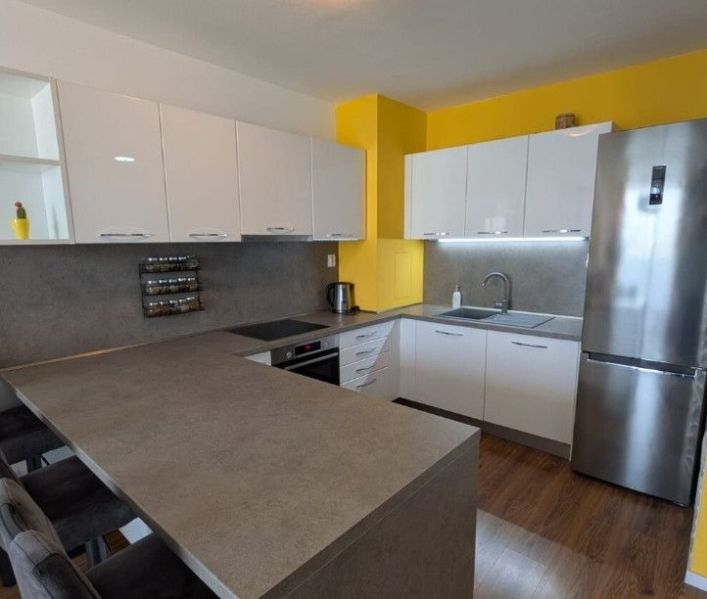 Moderný 2-izbový byt s balkónom a výhľadom - Kuchyňa v 2-izbovom byte s bielymi skrinkami, žltou stenou a podlahou s dreveným dekorom.