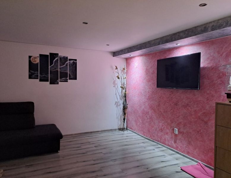 Rodinný dom Čachtice - Obývacia izba rodinného domu s podlahou s dreveným dekorom a televízorom na ružovej stene.