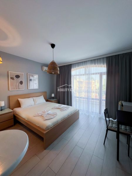 Moderná garsónka (41 m²) s výhľadom na bazén – Rezort ANTARES - Garsónka s podlahou v dekore dreva, veľkou posteľou a závesmi na okne.