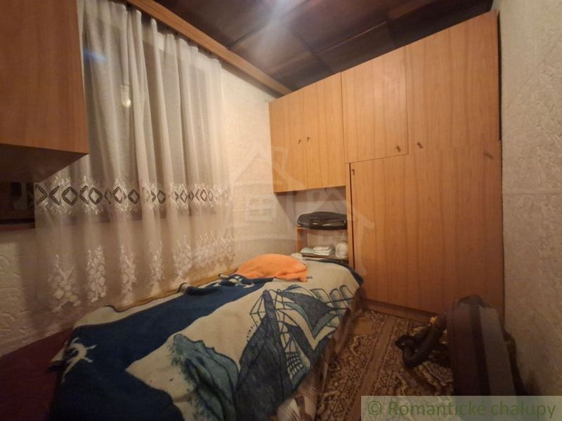 Poschodová chata pri lese blízko Dunaja v rekreačnej oblasti Kováčovsk - Interiér chaty s posteľou, skrinkami a vzorovaným kobercom, záclona na okne.
