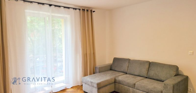 Prenájom 2 izb. bytu Račianske mýto, Bratislava - Obývacia izba so sivou sedačkou, veľkým oknom a závesmi v 2-izbovom byte.