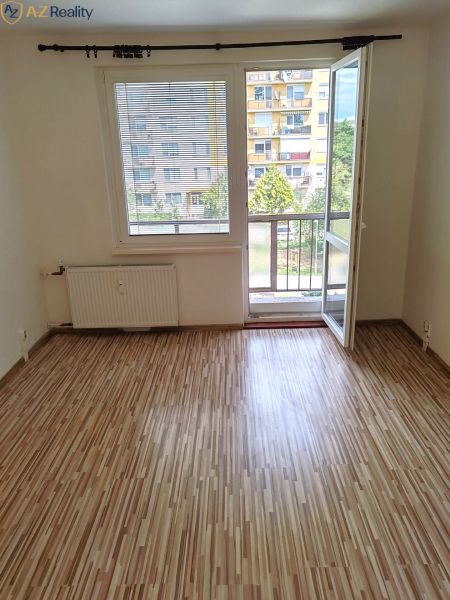 1 izbový byt s balkónom na prenájom  - Izba s podlahou s dreveným dekorom, balkónovými dverami a výhľadom na sídlisko.