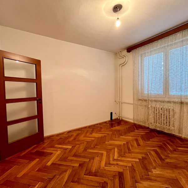 3-izbový byt s veľkou loggiou - Izba v 3-izbovom byte s podlahou s dreveným dekorom a oknom s bielou záclonou.