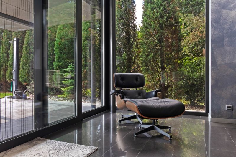 ARCHITEKTÚRA TICHA A MODERNÝCH TECHNOLÓGIÍ - SMART VILLA V OBJATÍ LESA - Kožené kreslo pri veľkých preskleniach s výhľadom na zeleň v rodinnom dome.