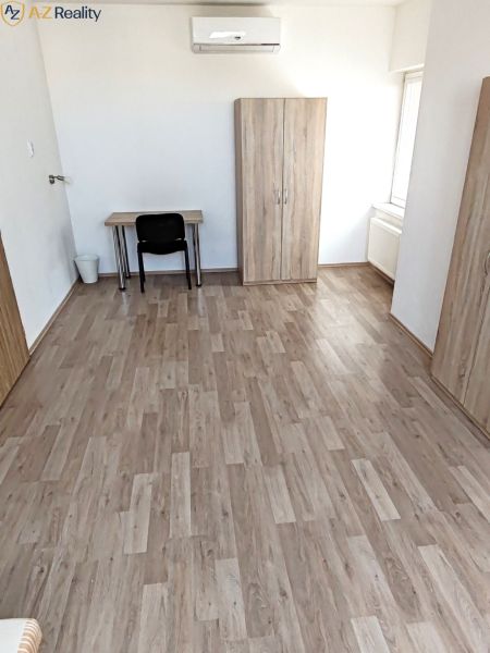 Väčší 1 izbový byt na prenájom v Komárne - Izba v 1-izbovom byte s podlahou s dreveným dekorom, stolom a skriňou.