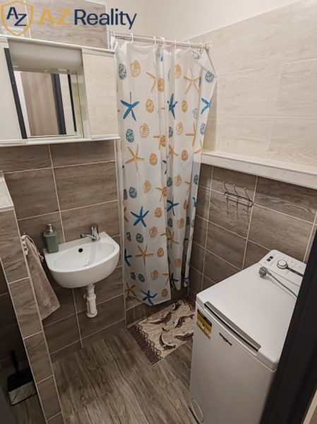 Pekný 1 izbový byt s balkónom na prenájom - Kúpeľňa v 1-izbovom byte s umývadlom, sprchou, práčkou a vzorovaným závesom.