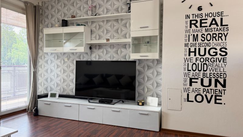 BÝVANIE V CENTRE MESTA: 3-IZBOVÝ BYT S LODŽIOU, TERASOU A GARÁŽOVÝM ST - Obývacia izba v 3-izbovom byte s televízorom, dekoračnou tapetou a podlahou s dreveným dekorom.