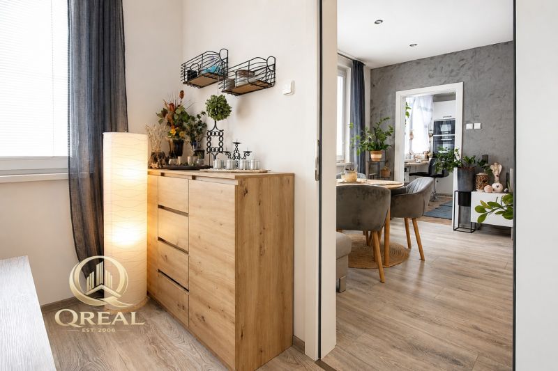 **QREAL**Moderné bývanie bez starostí**Len sa nasťahovať** - Interiér 2-izbového bytu s podlahou s dreveným dekorom, štýlovým nábytkom a rastlinami.