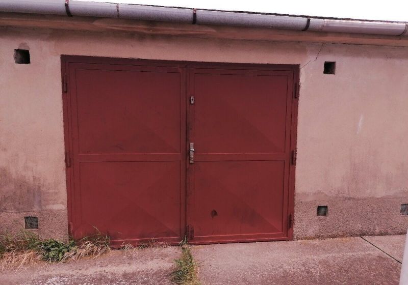 PREDAJ - Veľká murovaná garáž aj s pozemkom na ul. Rušňová - Rača - Zatvorené garážové dvere v Bratislave-Rači s opotrebovanými stenami a chodníkom.