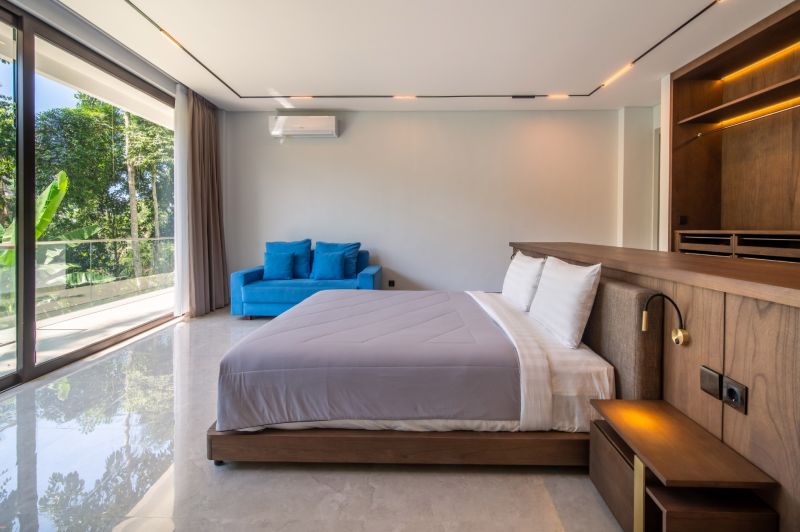 I Na predaj Luxusná vila „Tomma“ v Ubud, Bali - Spálňa s veľkými oknami, modrým gaučom a podlahou s dreveným dekorom vo vile.