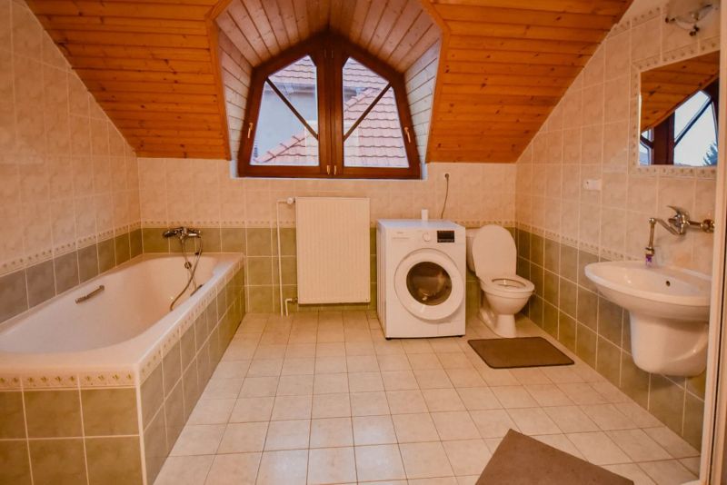 I Dom s wellness, bazénom a hotovým biznis potenciálom - Kúpeľňa s vaňou, práčkou a toaletou v rodinnom dome s dreveným stropom.