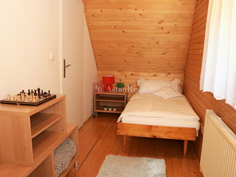 Chalupa Tále, okr.Brezno, pozemok 680m2,  - Jednolôžková izba s podlahou s dreveným dekorom, šachovnicou na stolíku a oknom s bielou záclonou.