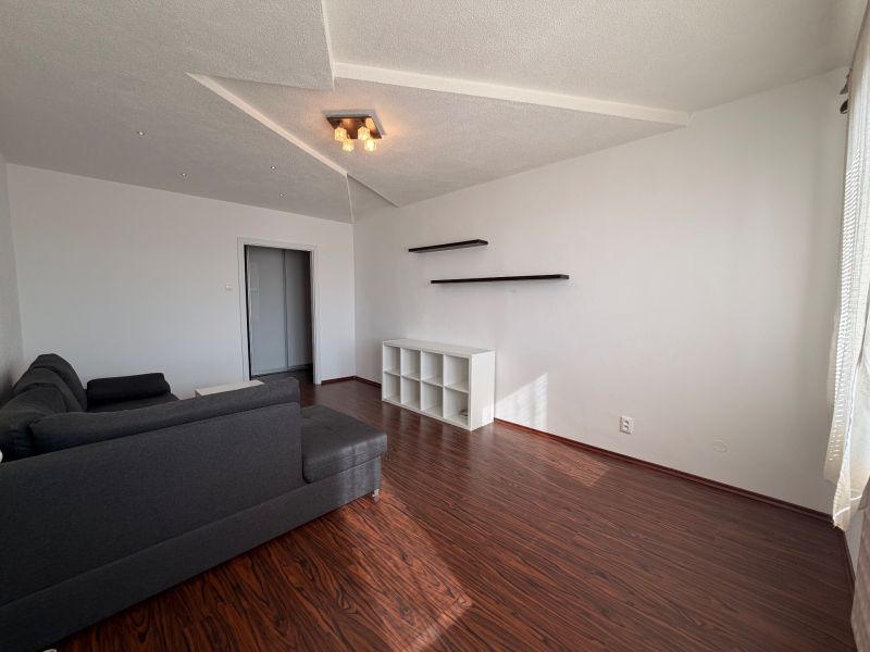 NOVINKA!!! Priestranný 3-izbový byt, 72 m² | Karpatská, Sásová - Obývačka v 3-izbovom byte s podlahou s dreveným dekorom, sivou sedačkou a poličkami.