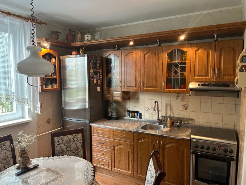NA PREDAJ PRIESTRANNÝ 3-IZBOVÝ BYT | 67 m² | LODŽIA | 4. POSCHODIE - Kuchyňa v 3-izbovom byte s dreveným nábytkom, dekoratívnou záclonou a jedálenským stolom.