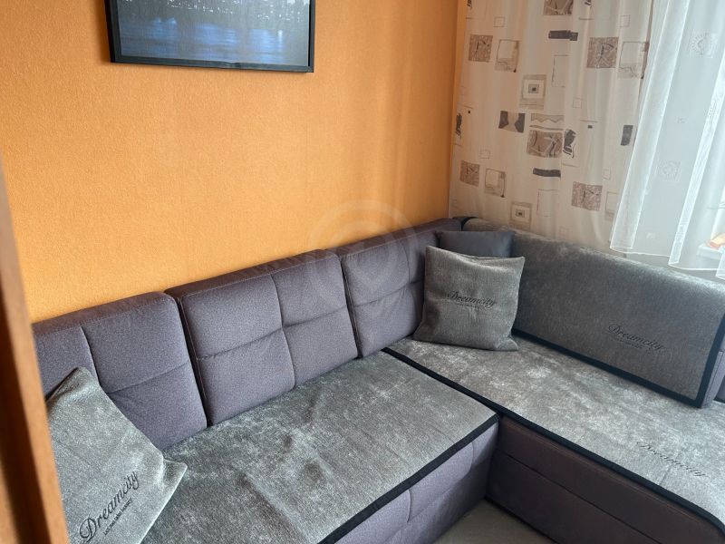 NA PREDAJ PRIESTRANNÝ 3-IZBOVÝ BYT | 67 m² | LODŽIA | 4. POSCHODIE - Šedá sedačka v rohu obývačky v 3-izbovom byte s oranžovou stenou.