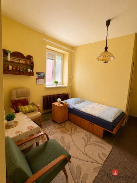 NA PREDAJ - Rodinný dom - Banská Bystrica - CENTRUM - Izba v rodinnom dome: posteľ, kreslá, stolík, koberec a okno so žaluziami.