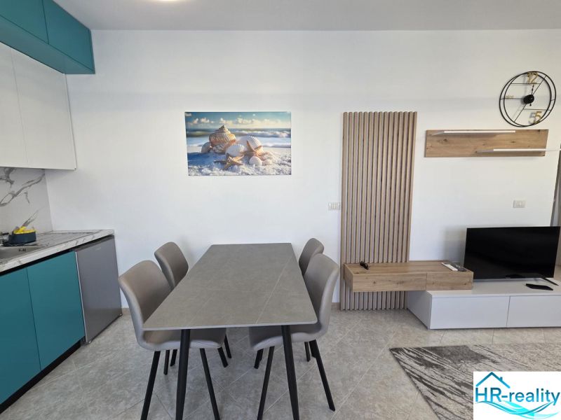 Albánsko, Durres - zariadený apartmán len 100m od mora a pláže - Svetlá kuchyňa v 2-izbovom byte so stolom, obrazom mušlí a televízorom.