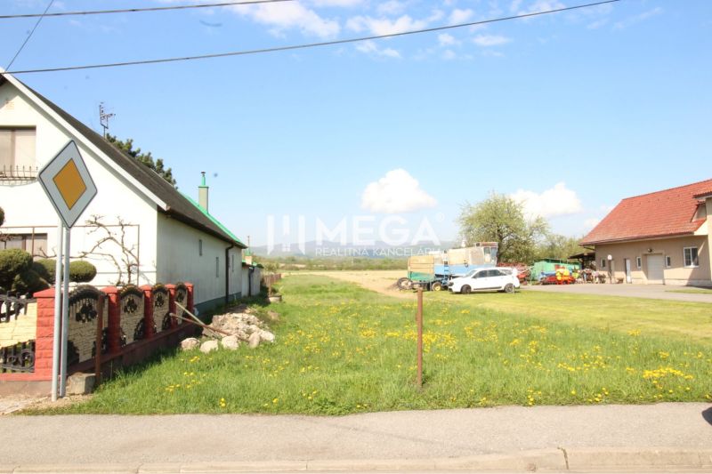 NA PREDAJ: Stavebný pozemok pri hlavnej ceste – Sačurov - Pozemky - bývanie v Sačurove s domami, trávnikom a cestnou značkou.