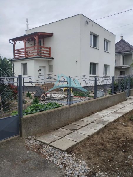 Váš nový rodinný prístav v obci Iža: Priestor, kľud a čerstvá úroda z  - Rodinný dom v meste Trnava s bielou fasádou a dreveným balkónom.