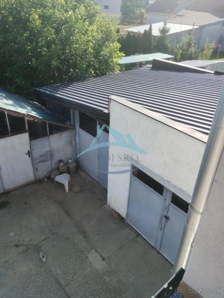 Váš nový rodinný prístav v obci Iža: Priestor, kľud a čerstvá úroda z  - Rodinný dom v Nitre s garážou, betónovým dvorom a zeleňou na pozadí.