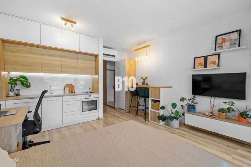 1-IZBOVÝ BYT S VEĽKOU 12 m² TERASOU | SLNEČNICE VILADOMY - 1-izbový byt s kuchyňou, pracovným stolom, TV a podlahou s dreveným dekorom.