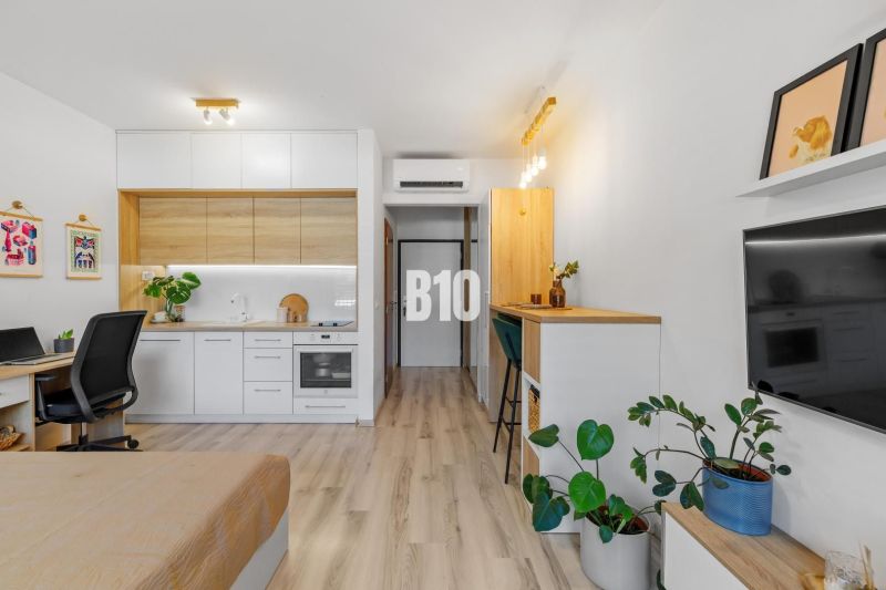 1-IZBOVÝ BYT S VEĽKOU 12 m² TERASOU | SLNEČNICE VILADOMY - Kompaktná kuchyňa s bielymi skrinkami a podlahou s dreveným dekorom v 1-izbovom byte.
