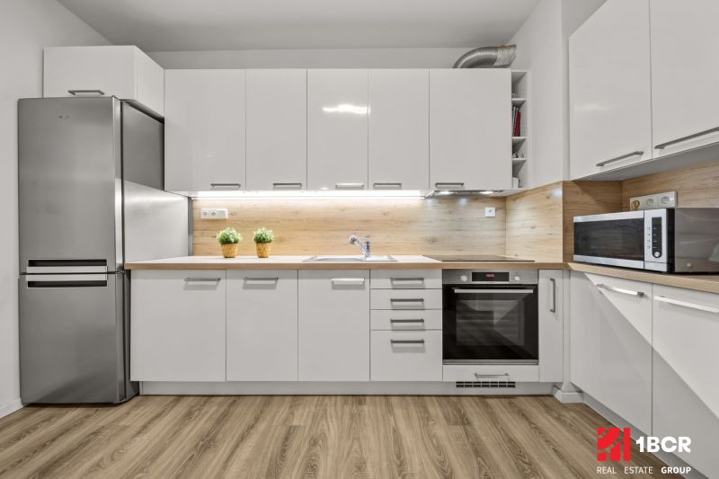 1.BCR | PRIESTRANNÝ 2-IZB. APARTMÁN V MAMAPAPA, PIVNICA, PARKING - Kuchyňa s bielymi skrinkami, nerezovou chladničkou a podlahou s dreveným dekorom v 2-izbovom byte.