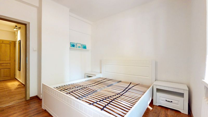 Priestranný 2i byt v obklopení zelene len pár krokov od OC Nivy | 53m² - Spálňa v 2-izbovom byte s podlahou s dreveným dekorom, bielou posteľou a nočným stolíkom.