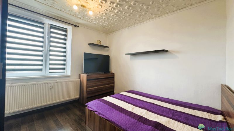 94,66 m², štyri izby a nikto nad hlavou. A Komenského má svoje výhody! - Spálňa v 4-izbovom byte s posteľou, televíziou, podlahou s dreveným dekorom a modernými žalúziami.