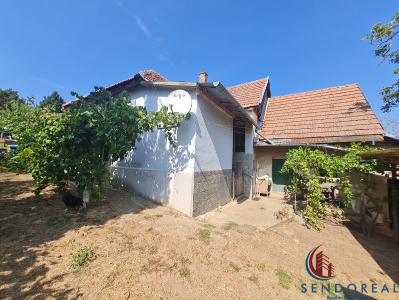 RD Otrokovce 1071m pozemok - Rodinný dom v Dolných Otrokovciach s bielou omietkou a škridlou, zelené rastliny na záhrade.