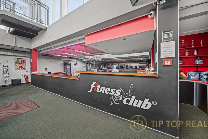 Na predaj PRIESTRANNÉ FITNESS CENTRUM na LUKRATÍVNEJ ADRESE RUŽINOVA - Recepcia fitness klubu s nápisom, červeným osvetlením a dekoratívnymi trofejami na zemi zeleného odtieňa.