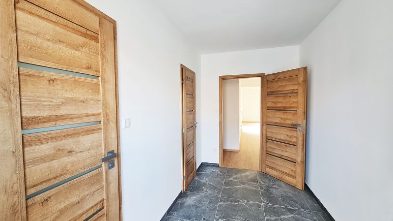 / Novostavba, moderný bungalov, dokončený na kľúč  - Chodba v rodinnom dome s dlažbou a drevenými dverami, biele steny.