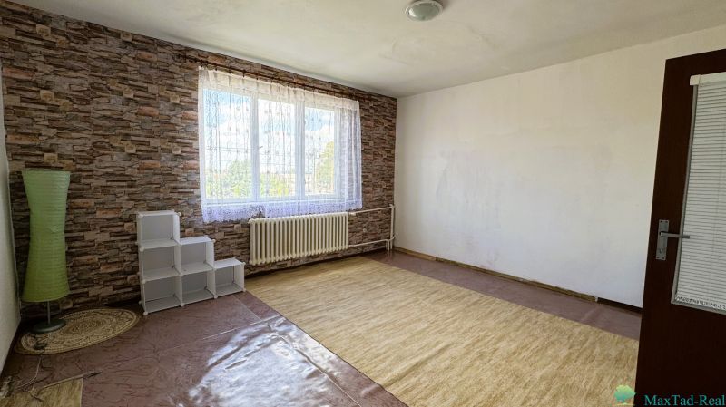 Dve generácie pod jednou strechou, Novosad, rodinný dom, 99.000€ - Izba s veľkým oknom, kobercom a tehlovým vzorom stien v rodinnom dome.