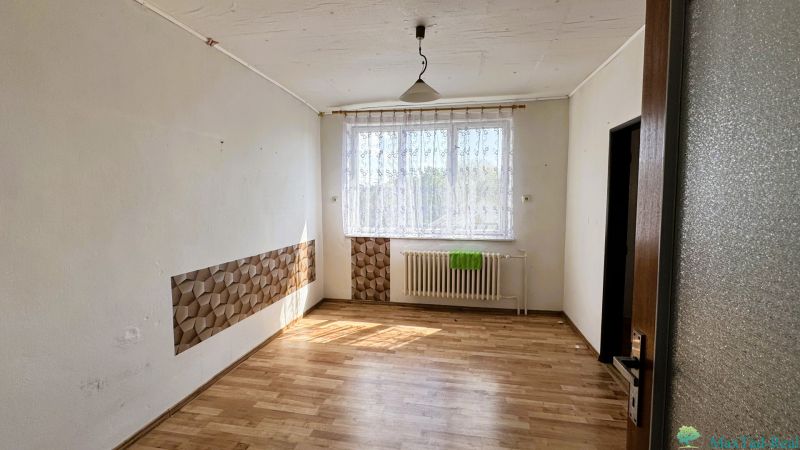 Dve generácie pod jednou strechou, Novosad, rodinný dom, 99.000€ - Izba v rodinnom dome s podlahou s dreveným dekorom, závesom a radiátorom pod oknom.